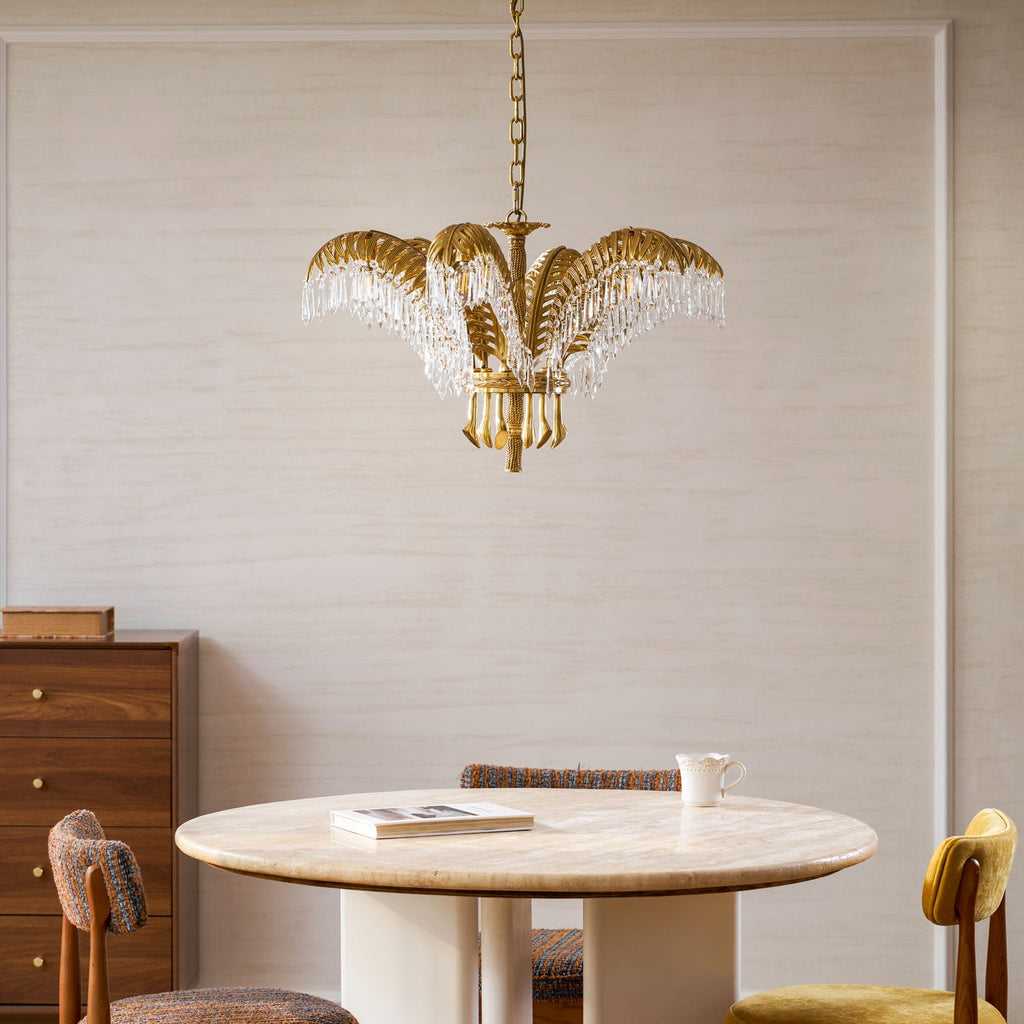 A small Clérachic brass chandelier hangs above a round stone-topped table with a book and a cup, in front of a pale wall.