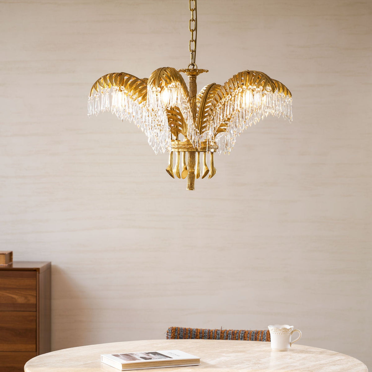 A Clérachic brass chandelier above a round table with a book, its crystal fringes lit warmly against a pale wall.