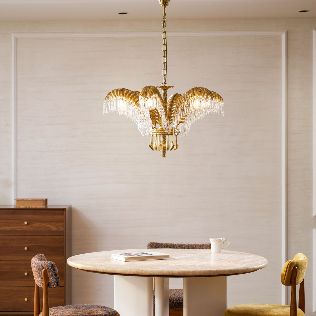 A Clérachic brass chandelier hung in a calm dining corner with a wooden chest of drawers, a round table and chairs.