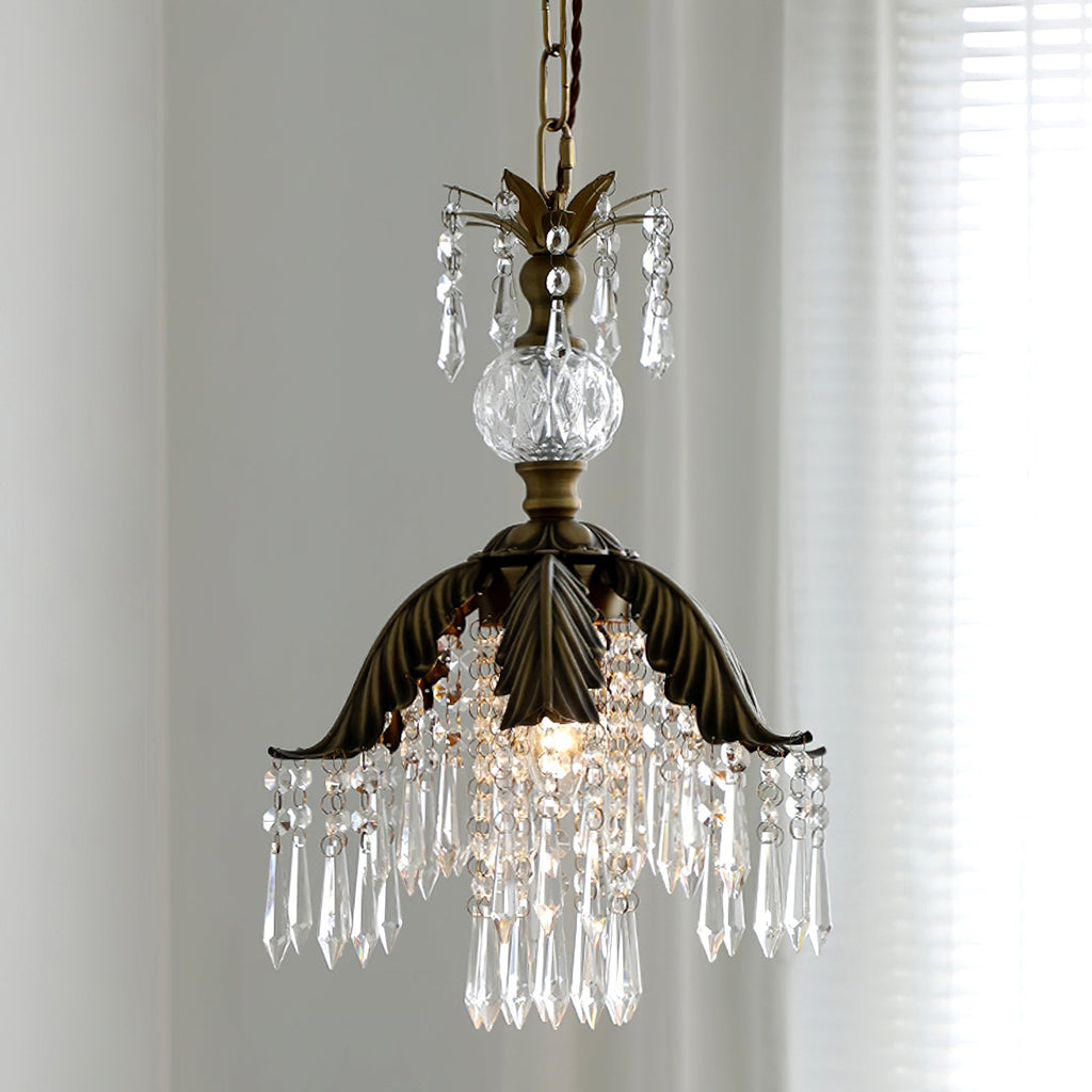 Low angle on a Clérachic pendant, its bronze palm-leaf shade and fringe of clear crystal drops lit from inside, with a sheer curtain behind.