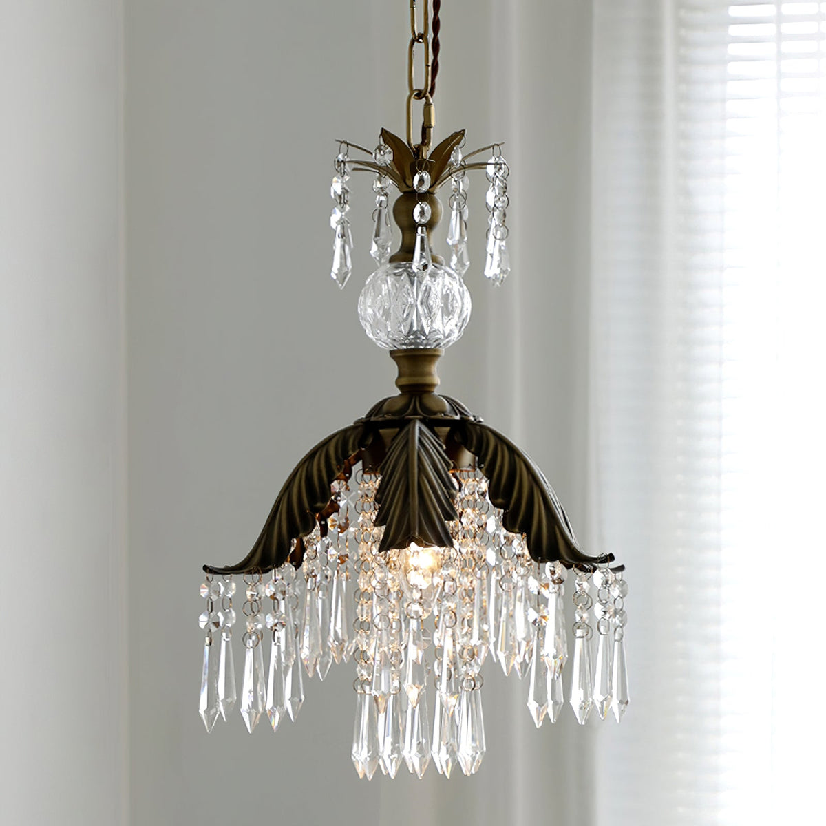 Low angle on a Clérachic pendant, its bronze palm-leaf shade and fringe of clear crystal drops lit from inside, with a sheer curtain behind.