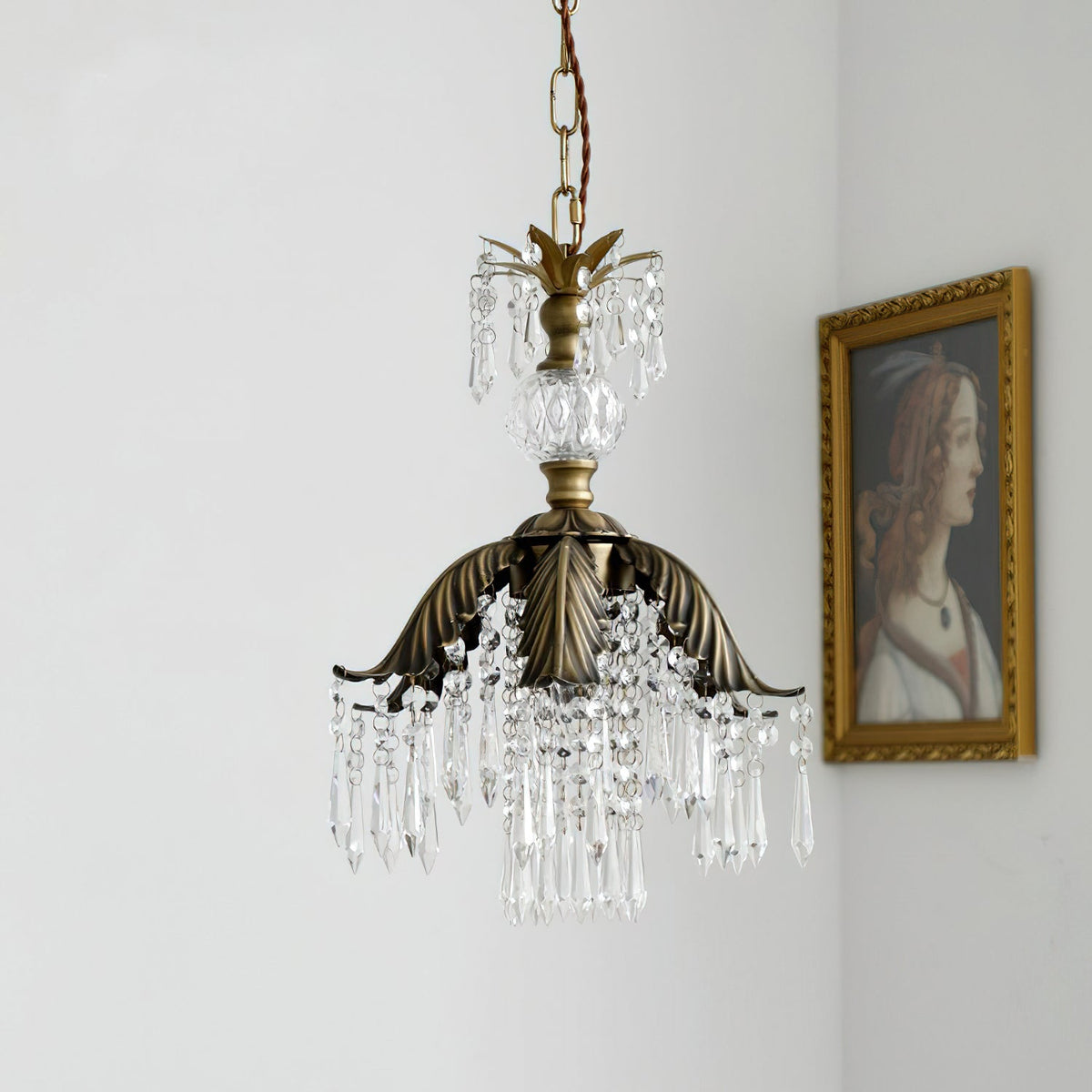 A Clérachic bronze-and-crystal pendant hangs unlit on a chain beside a gilt-framed portrait on a pale wall.