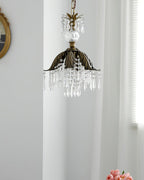 A Clérachic crystal pendant with a bronze leaf-shaped shade hangs before a white curtain and a small round gilt mirror.