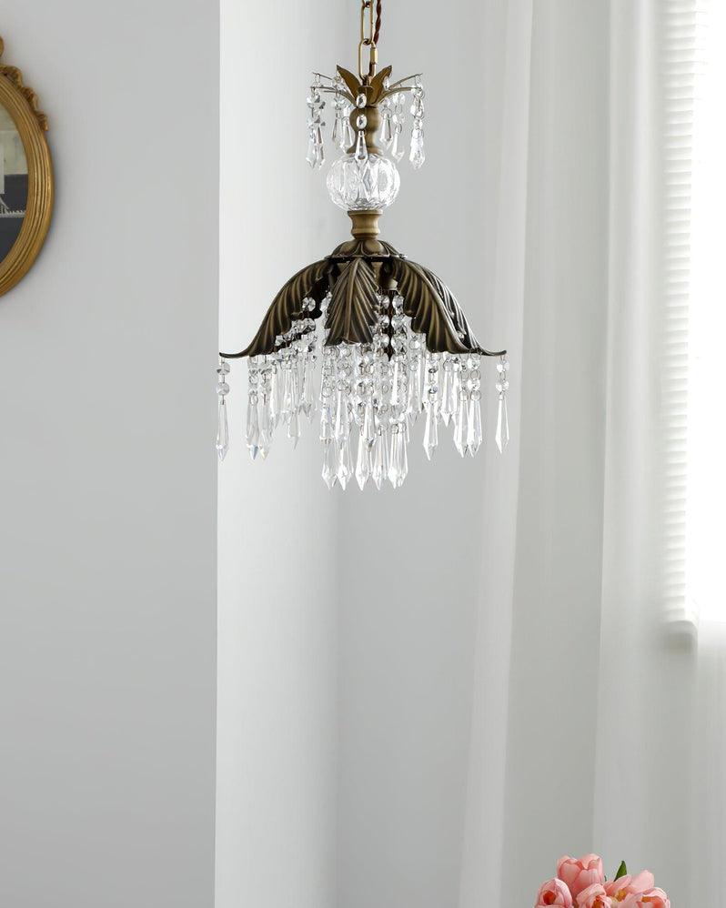 A Clérachic crystal pendant with a bronze leaf-shaped shade hangs before a white curtain and a small round gilt mirror.