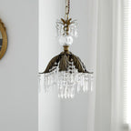 Clérachic pendant with a bronze palm-leaf shade and long crystal drops hangs unlit in front of a sheer curtain.