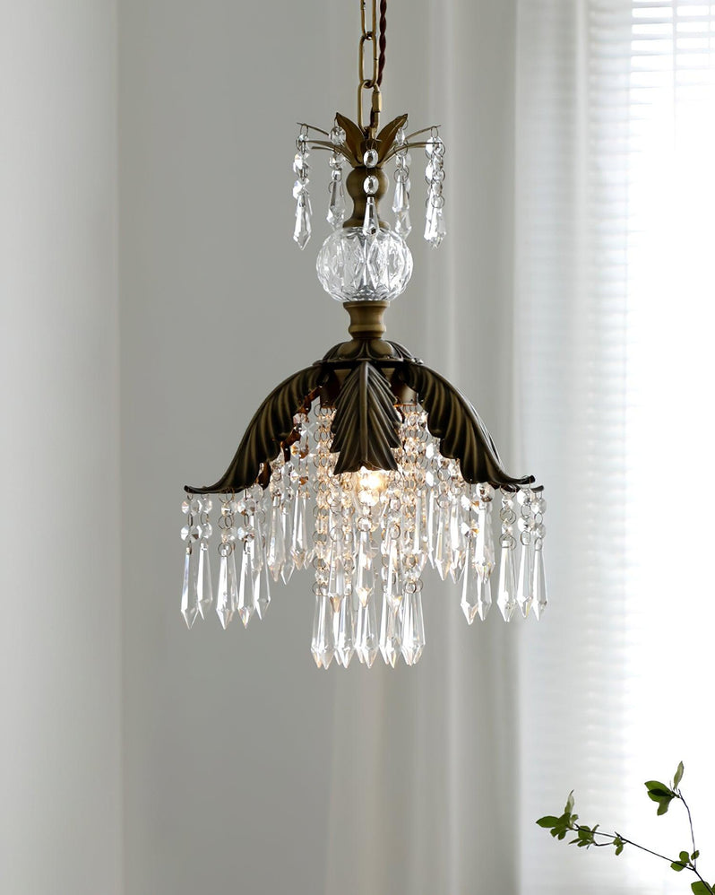 A lit Clérachic crystal pendant with a bronze palm-leaf shade hangs before a sheer curtain, with a green sprig at the corner.