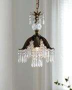 A lit Clérachic crystal pendant with a bronze palm-leaf shade hangs before a sheer curtain, with a green sprig at the corner.