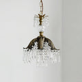 Clérachic pendant with a chain, glass finial, bronze palm-leaf shade and crystal fringe, hung against a pale grey wall.