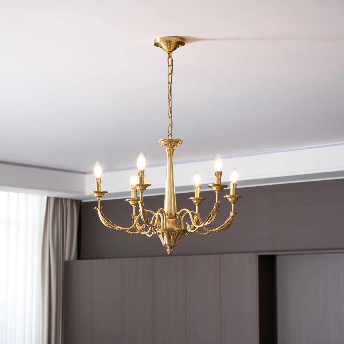 A brass Clérachic chandelier with six lit candle-style bulbs, hung from a chain below a recessed ceiling edge.