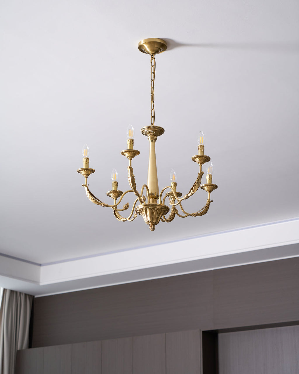 Hung from a ceiling canopy and chain above a dark panelled wall, this brass candle-style Clérachic chandelier is unlit.