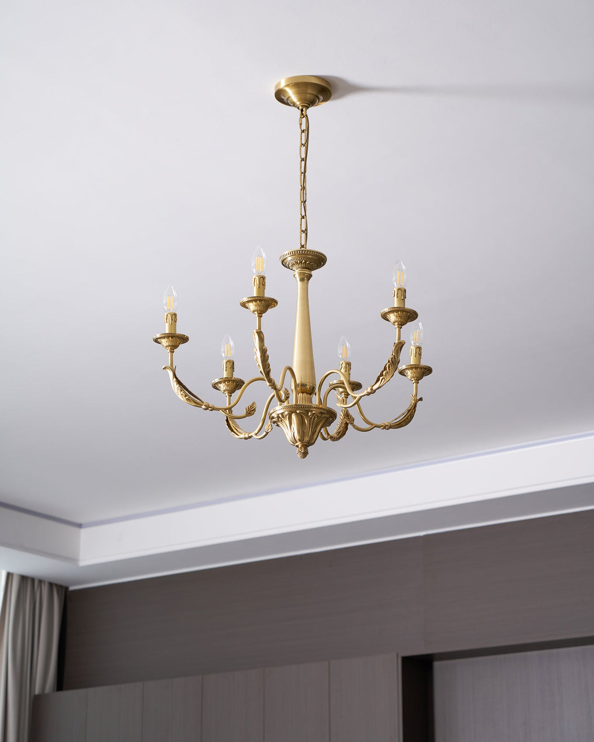 Hung from a ceiling canopy and chain above a dark panelled wall, this brass candle-style Clérachic chandelier is unlit.