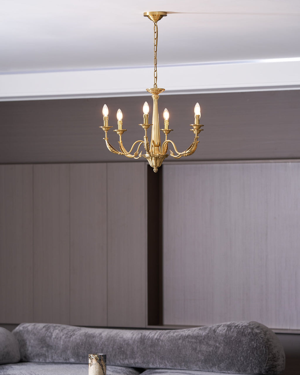 A brass candle-style Clérachic chandelier with six lit bulbs, seen from below, above a grey velvet sofa and dark wall.