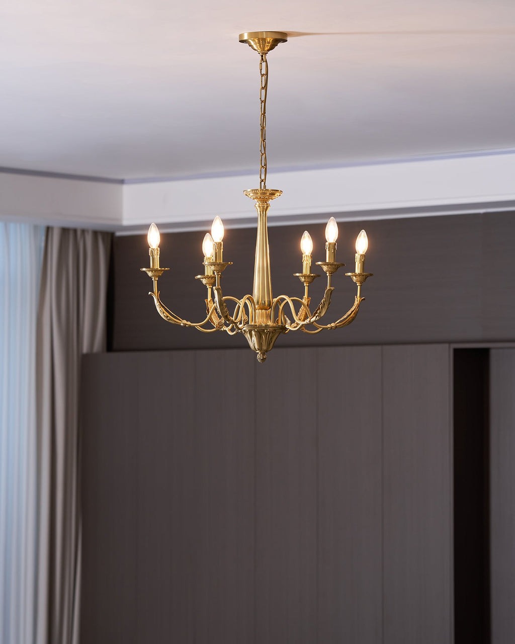 Six lit candle-style bulbs on this brass Clérachic chandelier, in front of a dark wooden wall and a pale curtain.