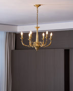 Six lit candle-style bulbs on this brass Clérachic chandelier, in front of a dark wooden wall and a pale curtain.