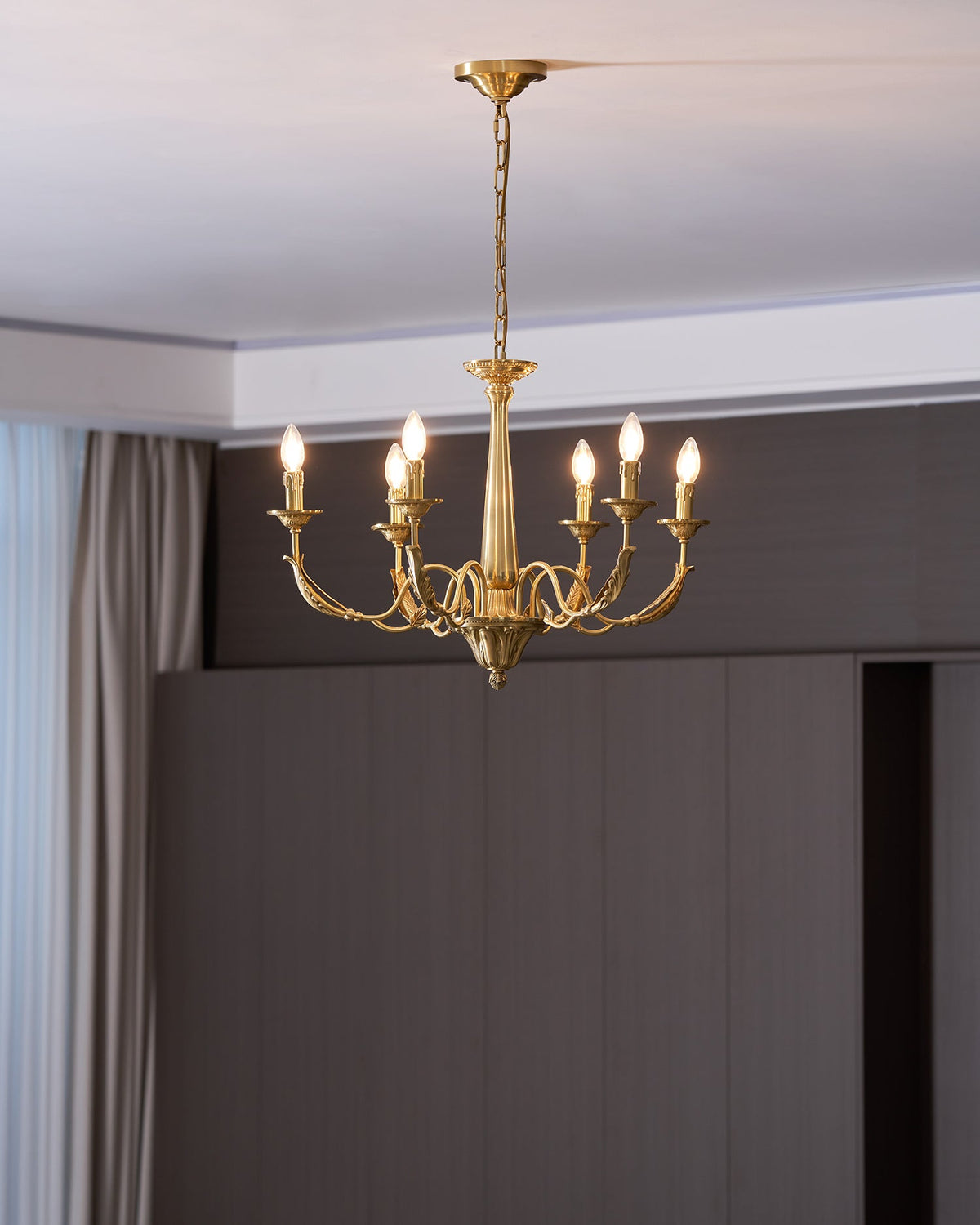 Six lit candle-style bulbs on this brass Clérachic chandelier, in front of a dark wooden wall and a pale curtain.