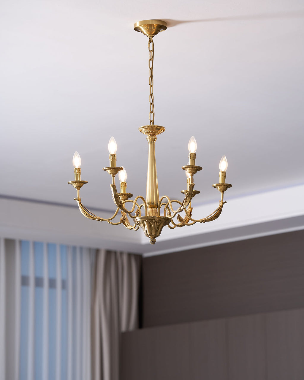 A brass candle-style Clérachic chandelier with six lit bulbs, hung from a long chain beside a curtain and a dark wall.