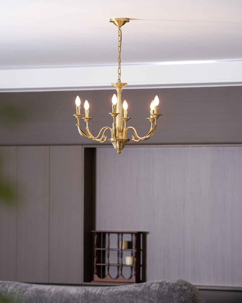 A brass candle-style Clérachic chandelier lit and seen from low down, with a grey sofa and a wine rack behind.
