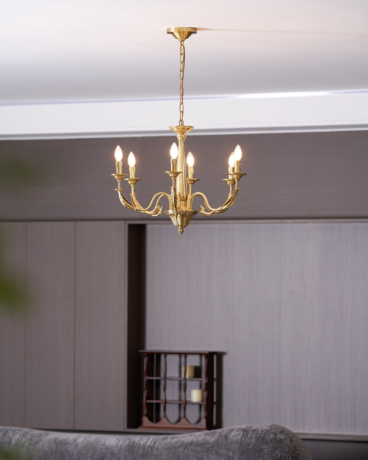 A brass candle-style Clérachic chandelier lit and seen from low down, with a grey sofa and a wine rack behind.