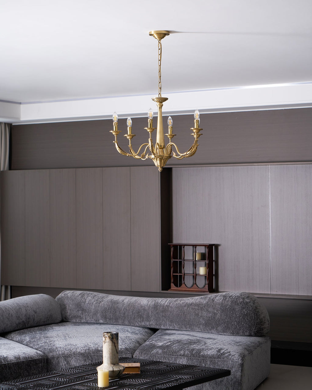 A brass candle-style Clérachic chandelier lit high in a living room with a grey sofa, a dark table and a wine rack.