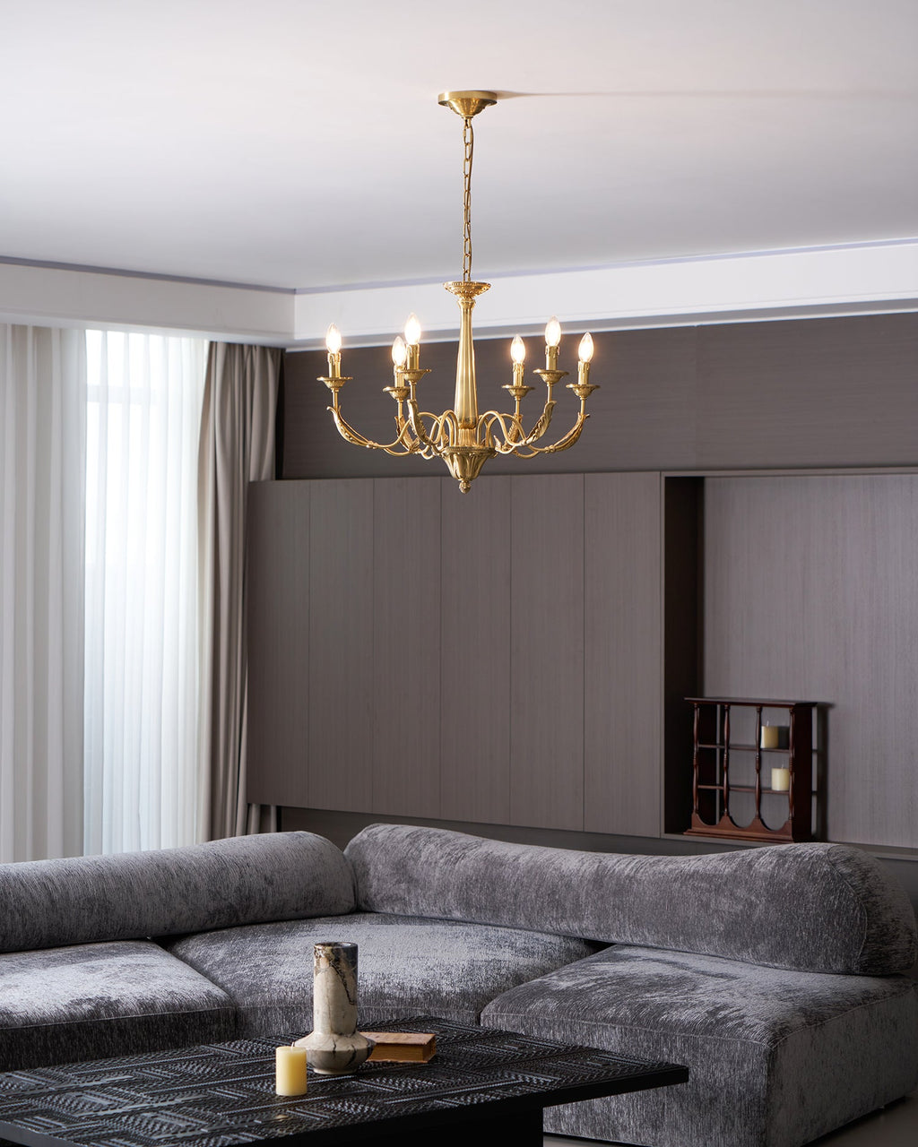 A brass candle-style Clérachic chandelier lit above a grey velvet sofa, with a curtain at the left and a dark wall behind.