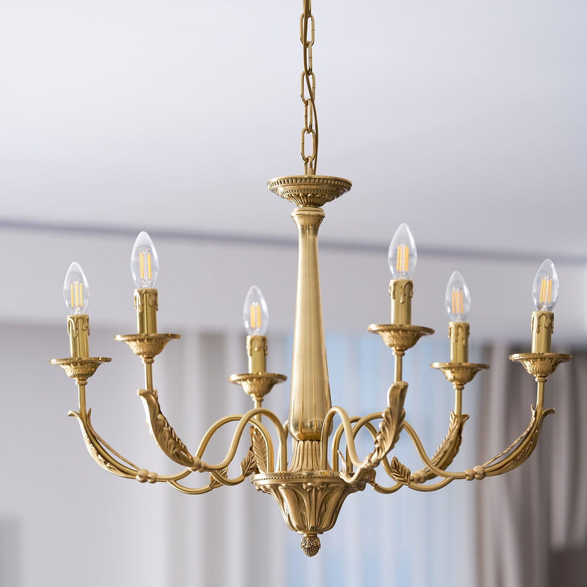 A brass Clérachic chandelier with six filament bulbs lit, hung on a chain, with a pale blurred room behind.