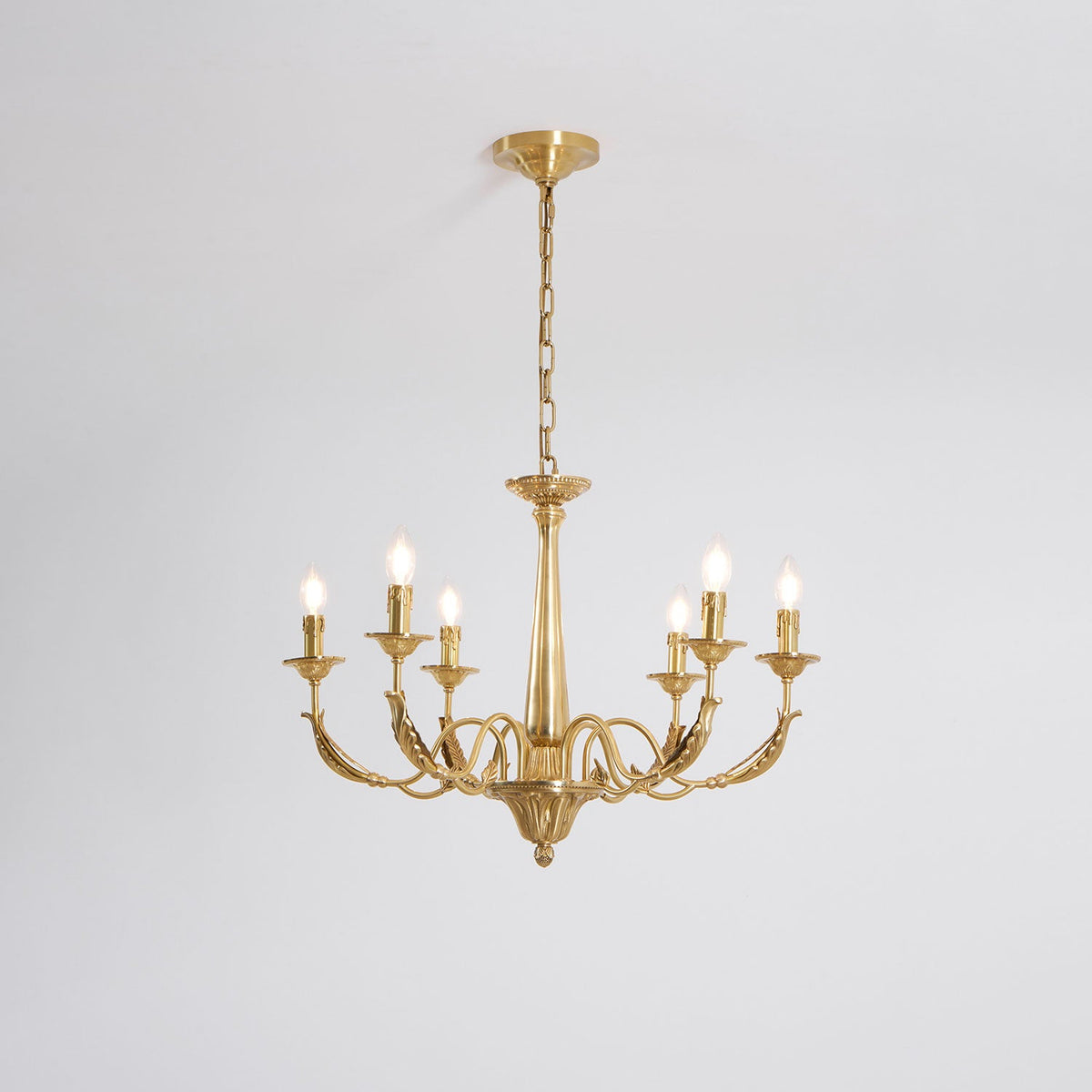 A brass candle-style Clérachic chandelier with six arms and tapered column, seen from slightly below on a pale grey backdrop.