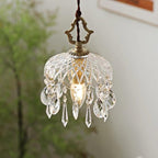 Close, softly lit view of a Clérachic pendant with a diamond-cut glass bowl, a candle bulb and clear crystal drops against green leaves.