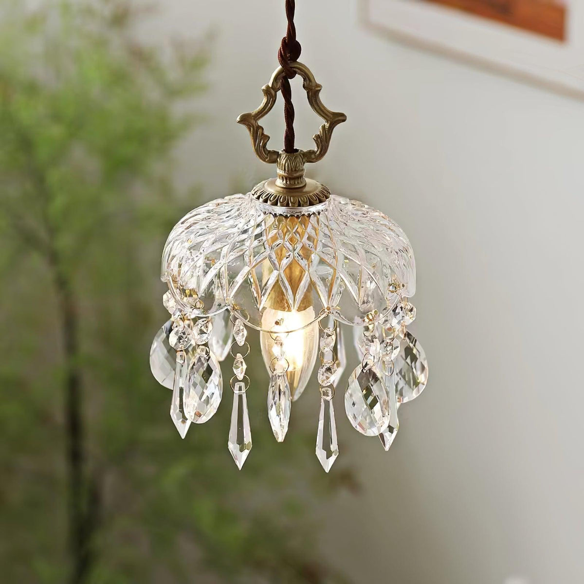 Close, softly lit view of a Clérachic pendant with a diamond-cut glass bowl, a candle bulb and clear crystal drops against green leaves.