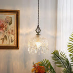 A lit Clérachic crystal-drop pendant beside a gilt-framed floral painting and a palm leaf, with sheer curtains glowing behind it.