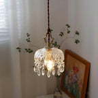 Clérachic cut-glass pendant lit beside a sheer curtain, with a bare green twig and a framed floral painting leaning on a table behind it.