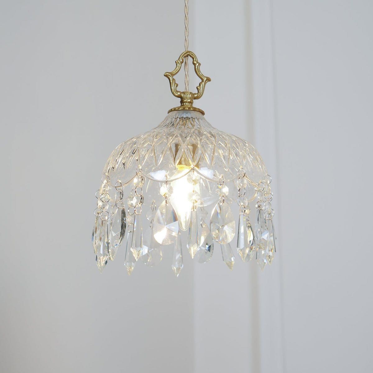 Clérachic crystal-drop pendant lit on a white wall, with a brass crown-shaped finial and a diamond-patterned glass bowl.