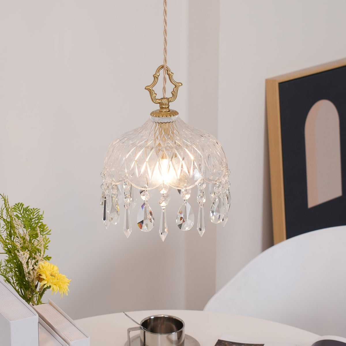 Clérachic glass pendant lit above a white table, with a yellow flower in a vase, a framed arch print and a steel cup nearby.