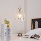 Clérachic crystal-drop pendant lit above a white table with stacked books, a steel cup and an open magazine, with a framed arch print behind.