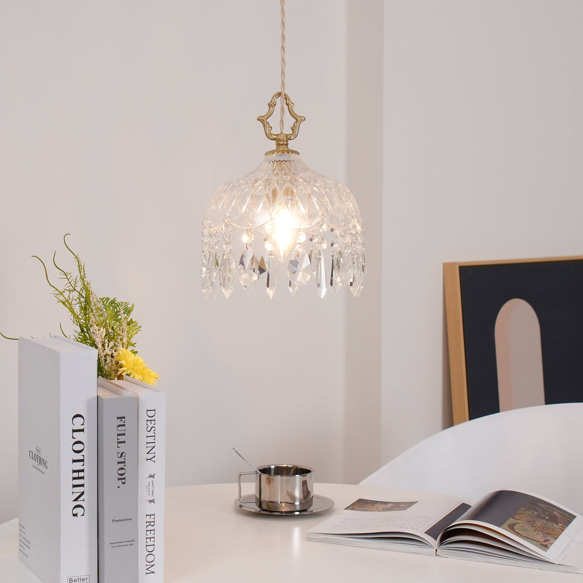 Clérachic crystal-drop pendant lit above a white table with stacked books, a steel cup and an open magazine, with a framed arch print behind.