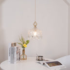 Clérachic crystal-drop pendant hung over a white round table with books, a gold vase of flowers and an open magazine below it.