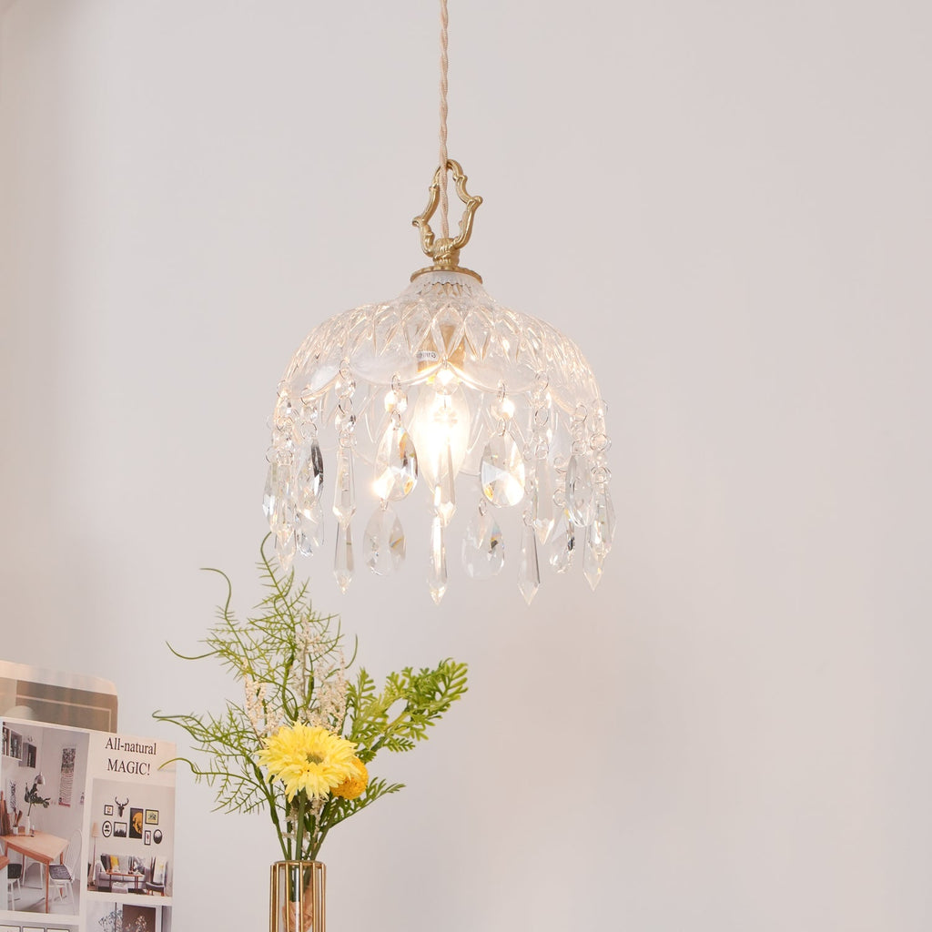 Clérachic faceted glass pendant lit against a blush wall, with a yellow flower and sprigs of greenery in a vase below.