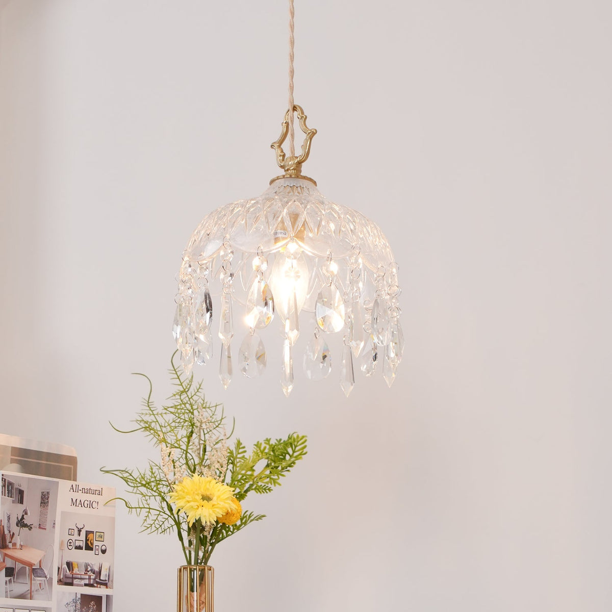 Clérachic faceted glass pendant lit against a blush wall, with a yellow flower and sprigs of greenery in a vase below.