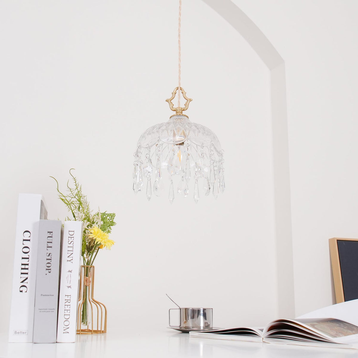 Clérachic crystal-drop pendant, pale and softly lit, on a white wall with a curved arch, above books and a vase with flowers.