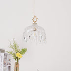 Clérachic crystal-drop pendant with a brass finial hung against a white wall, a vase of yellow flowers and stacked books below.
