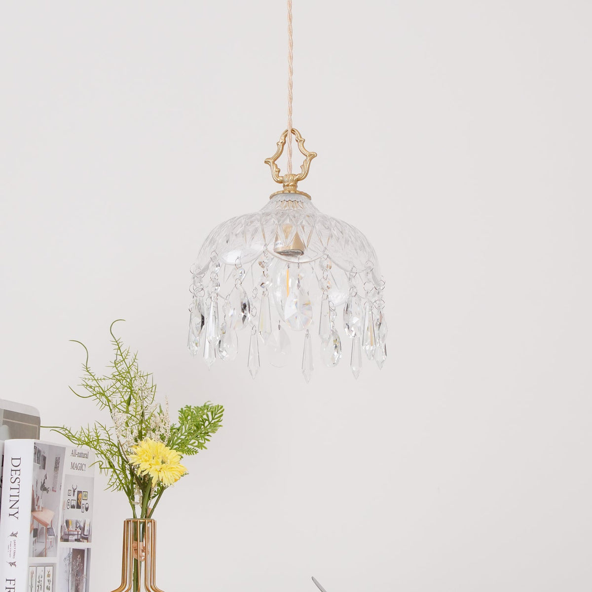 Clérachic crystal-drop pendant with a brass finial hung against a white wall, a vase of yellow flowers and stacked books below.