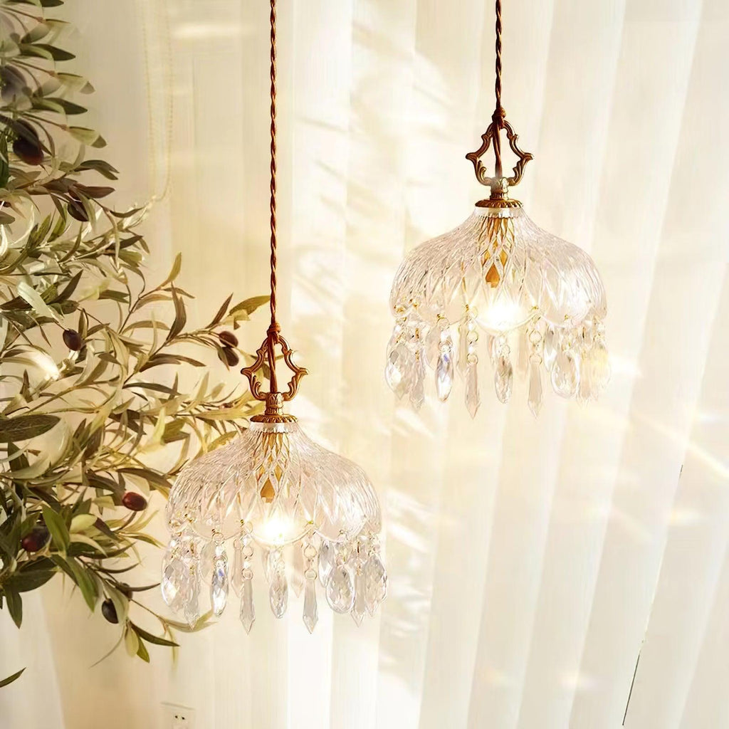 Two Clérachic crystal-drop pendants lit at different heights in front of cream curtains, with olive branches at the left.