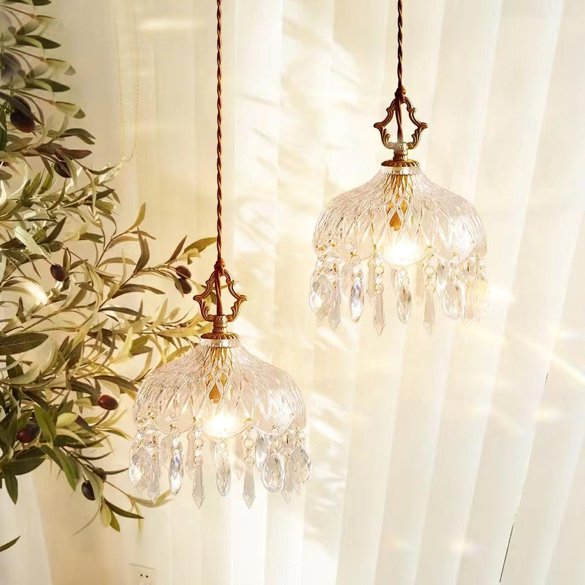 Two Clérachic crystal-drop pendants lit at different heights in front of cream curtains, with olive branches at the left.