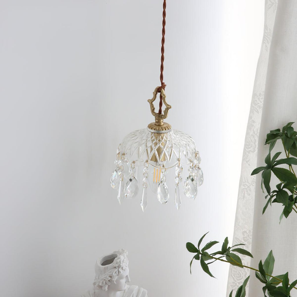 Clérachic crystal-drop pendant hung against a white curtain, lit softly, with green leaves at the lower right.