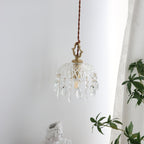 Clérachic crystal-drop pendant hung against a white curtain, lit softly, with green leaves at the lower right.