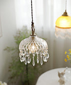 Clérachic crystal-drop pendant lit beside a sheer curtain, with an amber glass shade blurred in the upper right.