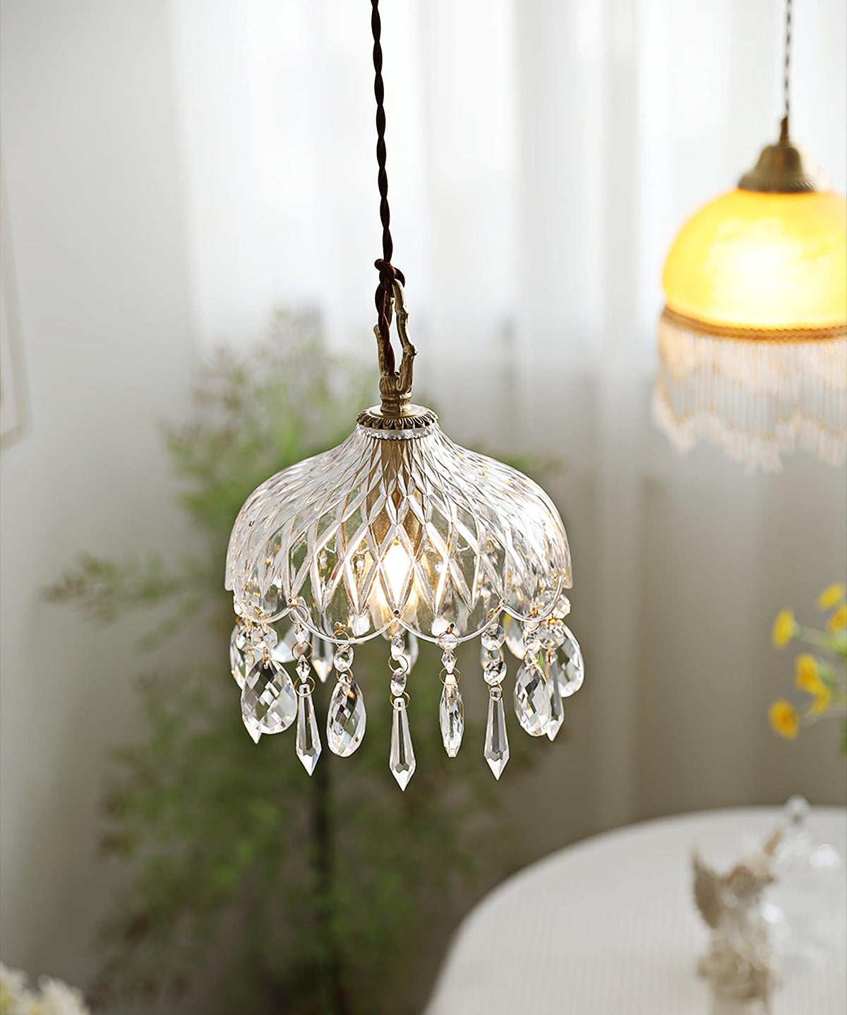 Clérachic crystal-drop pendant lit beside a sheer curtain, with an amber glass shade blurred in the upper right.