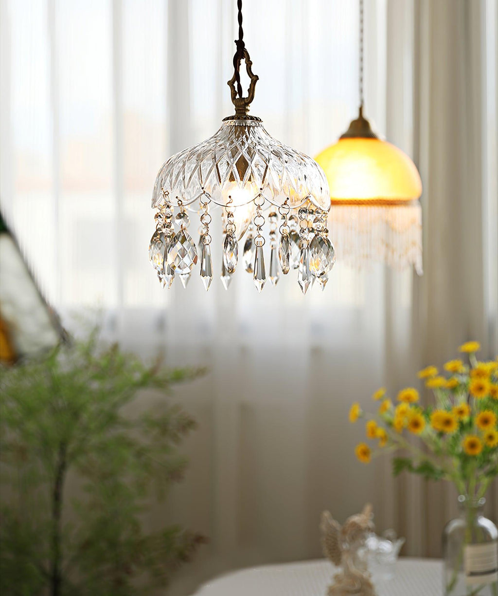 Clérachic crystal-drop pendant lit beside sheer curtains and yellow flowers, with an amber glass shade hung in the background.