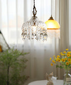 Clérachic crystal-drop pendant lit beside sheer curtains and yellow flowers, with an amber glass shade hung in the background.