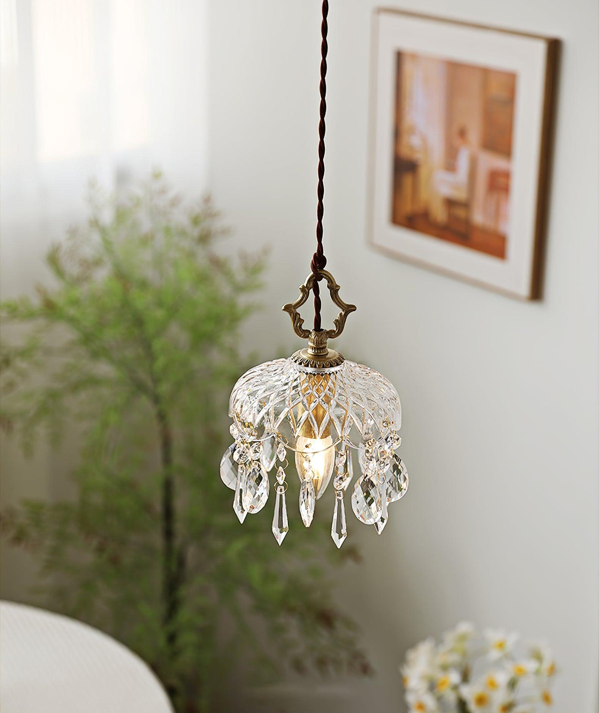 Clérachic crystal-drop pendant lit against a pale wall with a framed print, soft green foliage and white flowers below.