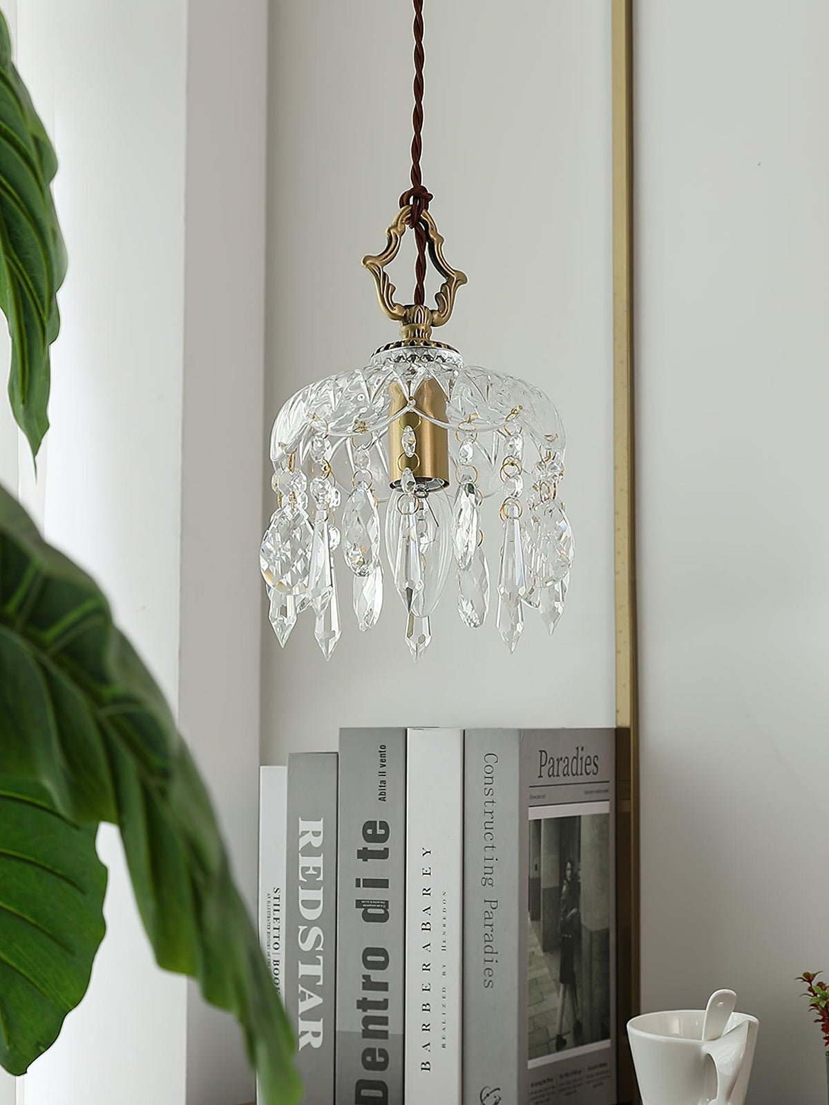 Clérachic crystal-drop pendant lit beside a tall green leaf and a stack of books on a shelf, with a wall mirror edge behind it.